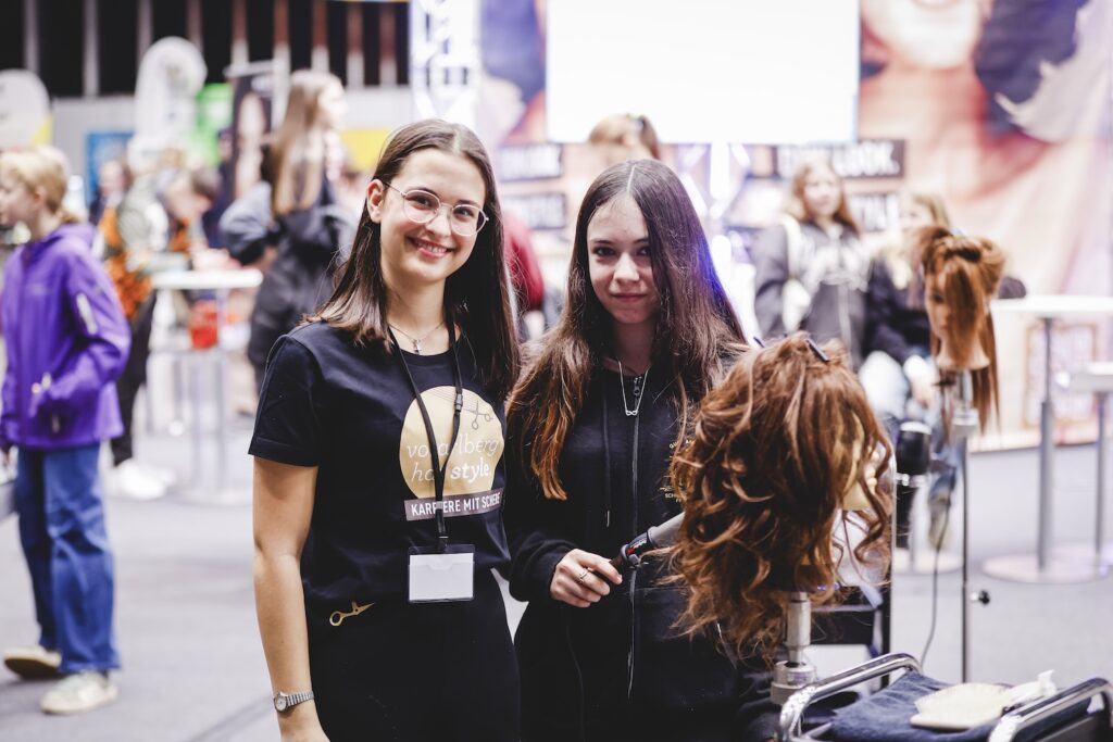Zwei junge Besucherinnen am Vorarlberg hat Style Stand, eine davon mit Übungskopf in der Hand, begleitet von einer Betreuerin.