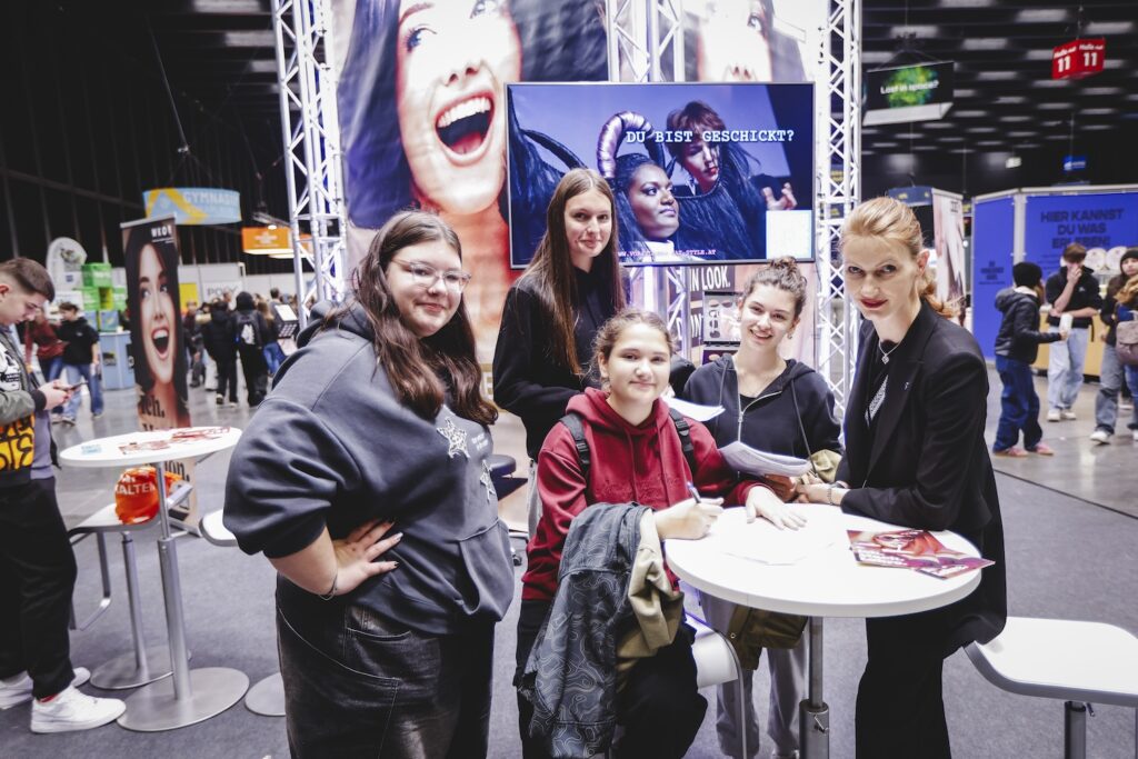 Jugendliche und eine Friseurin am Stand von Vorarlberg hat Style auf der i-Messe 2025, im Gespräch und mit Infomaterial am Tisch.